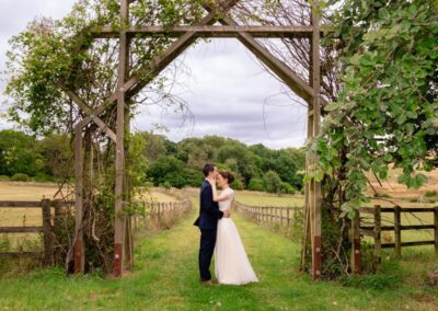 Sheepdrove Organic Farm in Berkshire. Bride and groom under entrance to lake pathway with beautiful countryside views.