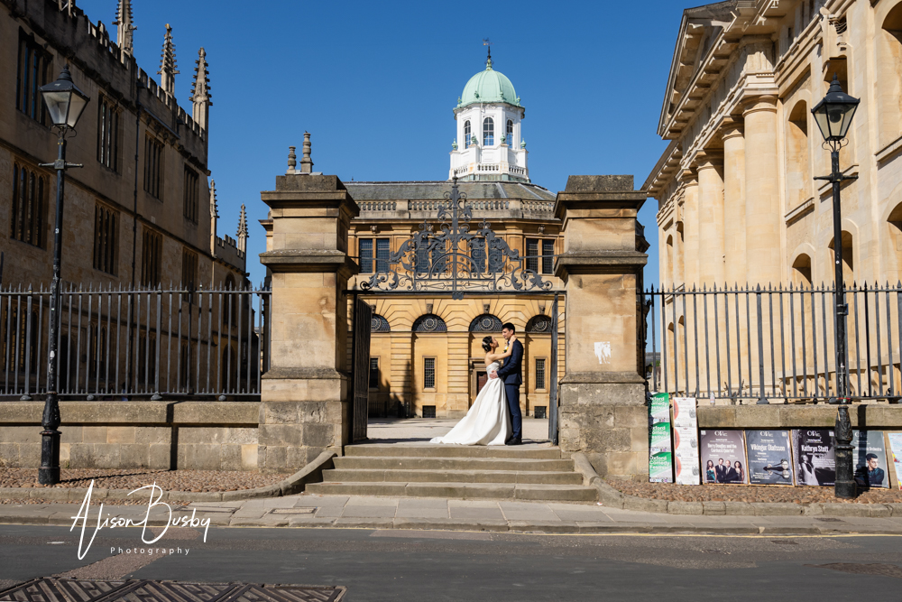 Bride and groom embrace at Great Gates of the Bodleian Library with the Sheldonian Theatre and Clarendon Building in background. Oxford. By Bodleian Library wedding photographer.