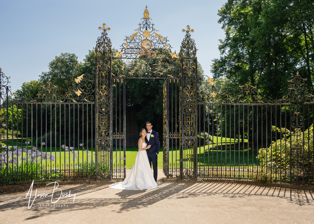 Wedding photos Oxford. Bride and groom at gate to the gardens of New College Oxford with blue sky and deep shadows in background.