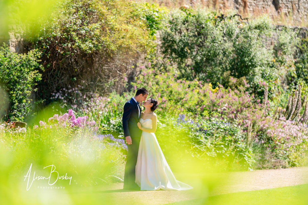 Bride and groom in gardens of New College Oxford with background sunlit flowers. Oxford wedding day.