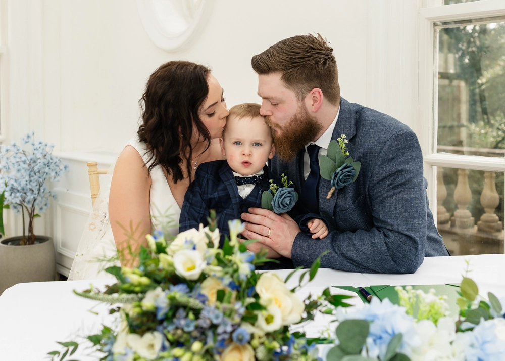 Bride and groom kissing small child on head after ceremony in conservatory. Hunton Park wedding photographer.