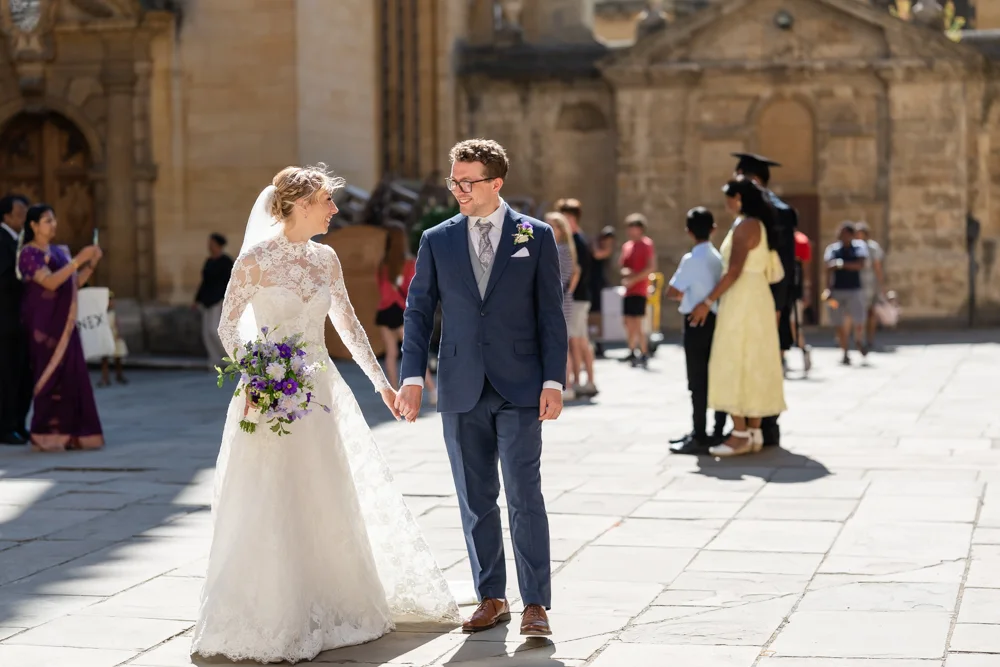 Oxford University wedding photographer. Bride and groom walking with Oxford graduates in background.